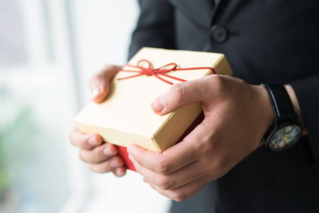 Close-up of a person wearing a dark suit and watch holding a small, cream-colored gift box tied with a thin red ribbon